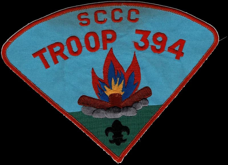 The Troop 394 neckerchief patch: a campfire on a blue sky above green ground, under the words SCCC Troop 394, with a fleur-de-lis below the fire.